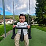 child, toddler, swing, playground, outdoor, grass, trees, sky, clouds, houses, greenery, sandals, pants, shirt, happy, smile, person, daylight, nature, fun