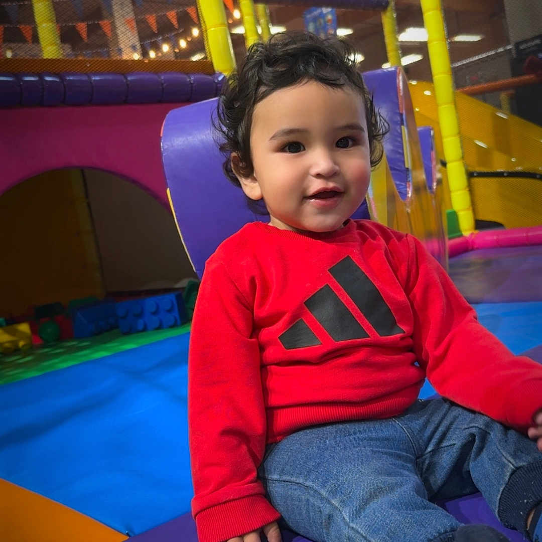 Lorenzo a rejoint le concours — aidez-le/la à gagner de superbes lots ! bright_colors, casual_clothing, child, climbing_structure, colorful_mats, curly_hair, footwear, happy, indoor_lighting, indoor_play_area, jeans, person, playground_equipment, playtime, red_sweatshirt, seated, smiling, soft_play, toddler, young_child