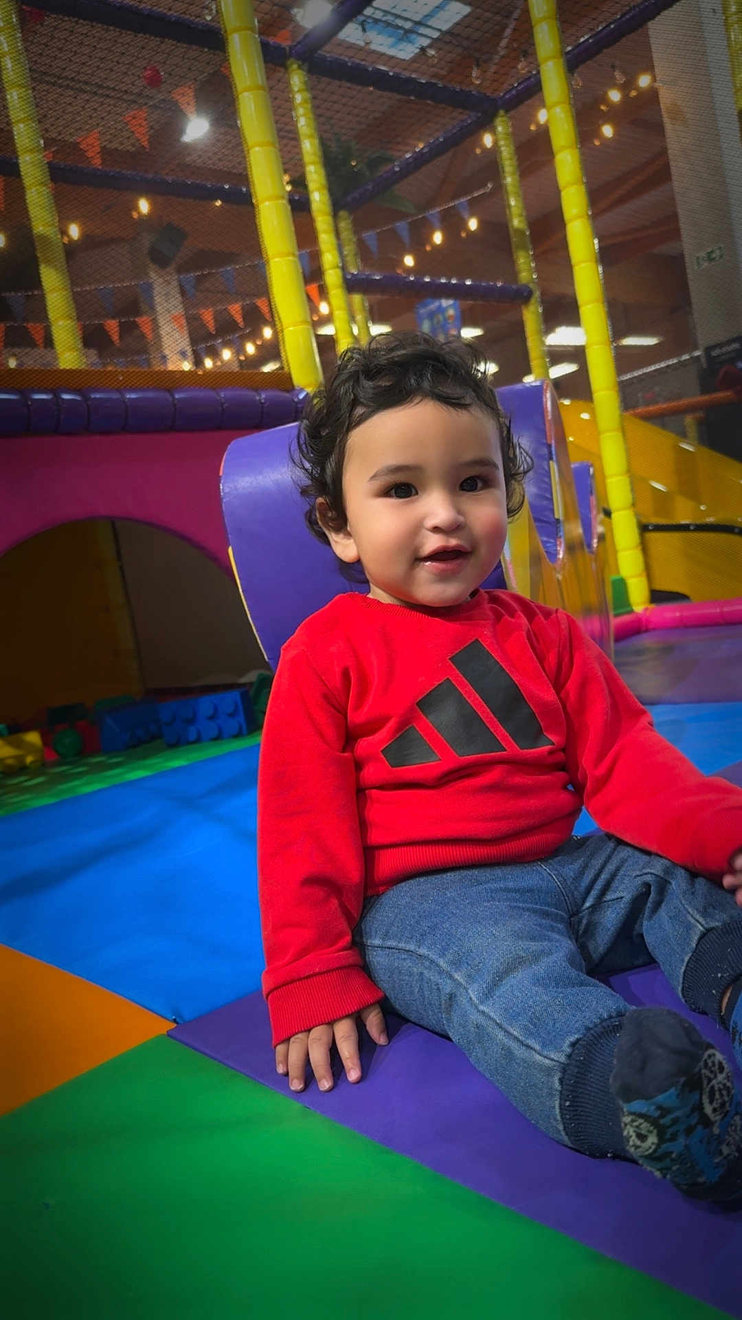 Lorenzo a rejoint le concours — aidez-le/la à gagner de superbes lots ! toddler, child, indoor_play_area, colorful_mats, red_sweatshirt, jeans, curly_hair, smiling, playground_equipment, soft_play, climbing_structure, happy, casual_clothing, playtime, young_child, person, footwear, seated, bright_colors, indoor_lighting