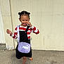 child, smiling, girl, hair_buns, striped_shirt, black_pinafore, purple_bag, sandals, outdoor, concrete_ground, beige_wall, accessories, happy, posing, casual_clothing, playful, daylight, person, fashion, cute