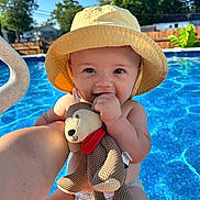 Vinny is registered to the contest to win money with this photo: baby, child, yellow_hat, plush_toy, teddy_bear, pool, water, sunlight, outdoor, summer, smile, happy, cute, diaper, hand, face, blue, sky, trees, fence