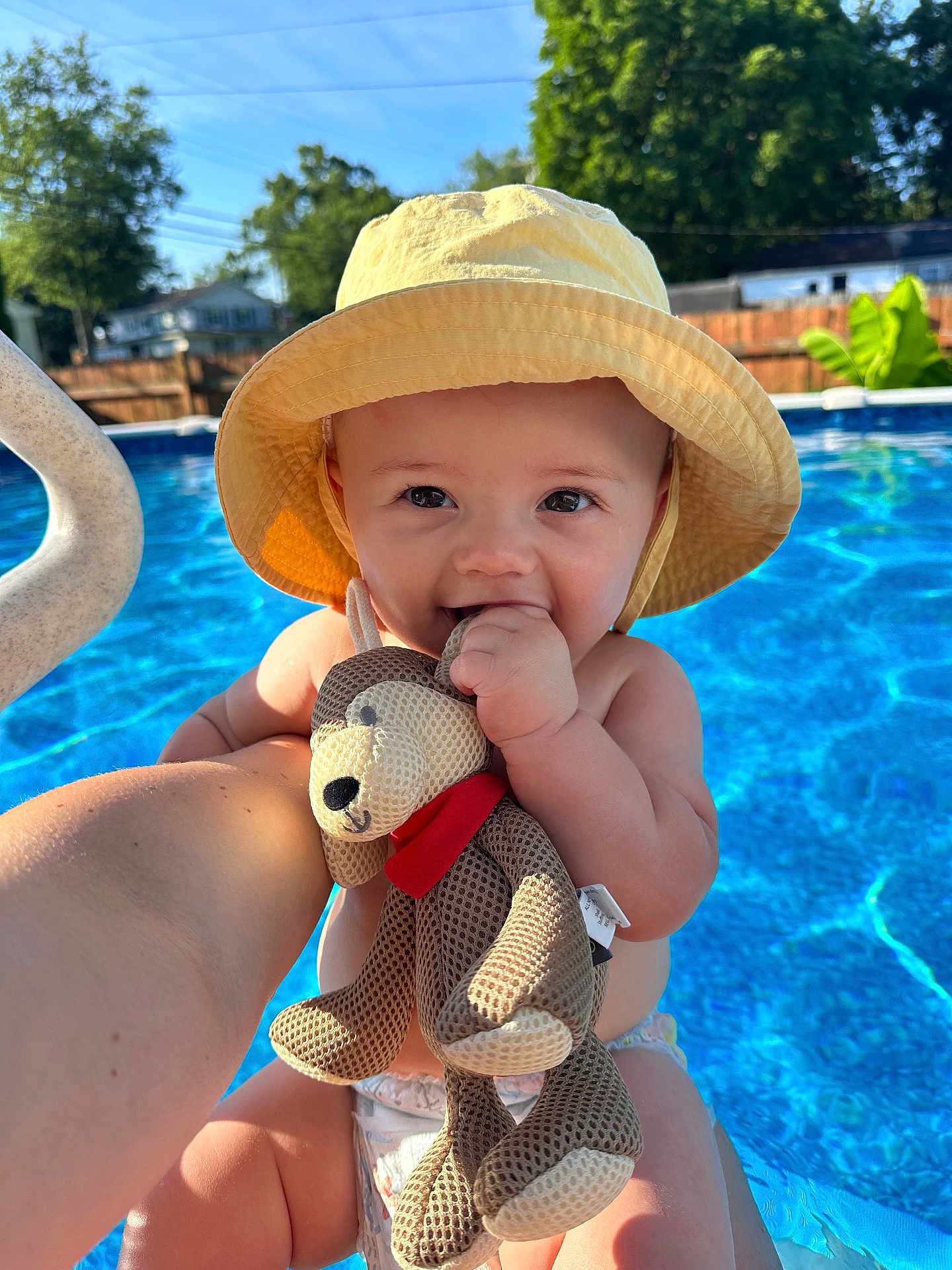 Vinny is registered to the contest to win money with this photo: baby, child, yellow_hat, plush_toy, teddy_bear, pool, water, sunlight, outdoor, summer, smile, happy, cute, diaper, hand, face, blue, sky, trees, fence