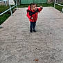 Logan participe au concours pour gagner de l'argent avec cette photo : child, boy, red_jacket, backpack, outdoor, concrete, path, green_gate, building, grass, fence, curious, playful, casual_clothing, shoes, daylight, sidewalk, urban, small_child, smiling
