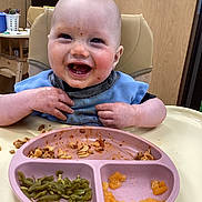 Liam is registered to the contest to win money with this photo: baby, bib, child, clothing, diced_peaches, face, feeding, food, green_beans, hands, happy, high_chair, indoor, meal, messy_face, pasta, person, plate, smile, table