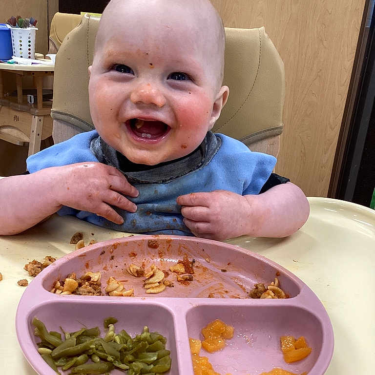 Liam is registered to the contest to win money with this photo: baby, bib, child, clothing, diced_peaches, face, feeding, food, green_beans, hands, happy, high_chair, indoor, meal, messy_face, pasta, person, plate, smile, table