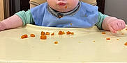 Liam is registered to the contest to win money with this photo: baby, child, high_chair, blue_bib, food, orange_food, face, cheeks, funny_face, table, tray, clothing, sitting, indoor, wooden_chair, expression, meal_time, cute, person, infant