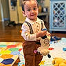 Jonathan is registered to the contest to win money with this photo: toddler, child, toy, plush_cow, brown_pants, suspenders, patterned_shirt, indoor, play_mat, colorful, wooden_floor, cozy_room, soft_lighting, standing, smiling, cute, person, hair_tuft, footwear, home