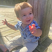Maverick is registered to the contest to win money with this photo: toddler, child, smiling, outdoor, sunlight, wooden_deck, dinosaur_shirt, happy, sitting, barefoot, portrait, person, nature, daylight, cute, casual_clothing, baby, expression, young, playful