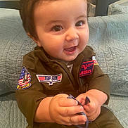Maverick is registered to the contest to win money with this photo: baby, child, clothing, couch, cozy, cute, face, hands, happy, indoor, infant, patches, person, pilot_jumpsuit, portrait, quilt, seated, smiling, sunglasses, young_child