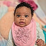 baby, child, closeup, curly_hair, cute, face, floral_design, hand, indoors, infant, pastel_colors, person, pink_bib, portrait, skin, smiling, soft_background, white_clothing, wide_eyes, young