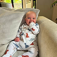 Elliot is registered to the contest to win money with this photo: baby, onesie, dinosaur_pattern, couch, cushion, indoor, smiling, happy, child, infant, hand_in_mouth, natural_light, window, home, furniture, cozy, person, seated, soft_texture, cute
