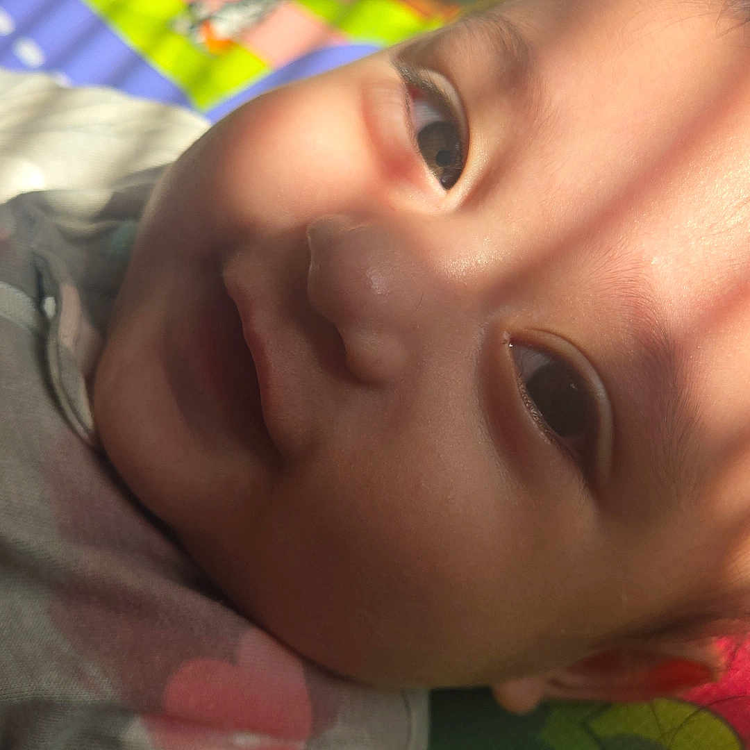 Samuel is registered to the contest to win money with this photo: baby, child, face, close_up, play_mat, colorful, sunlight, shadow, smile, infant, skin, eyes, clothing, indoors, portrait, cute, young, soft_light, relaxing, happy