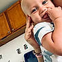 baby, child, blue_eyes, hand, fingers, kitchen, cabinet, wall, photographs, person, closeup, infant, clothing, sleeve, adult_hand, holding, smiling, cute, indoor, playful