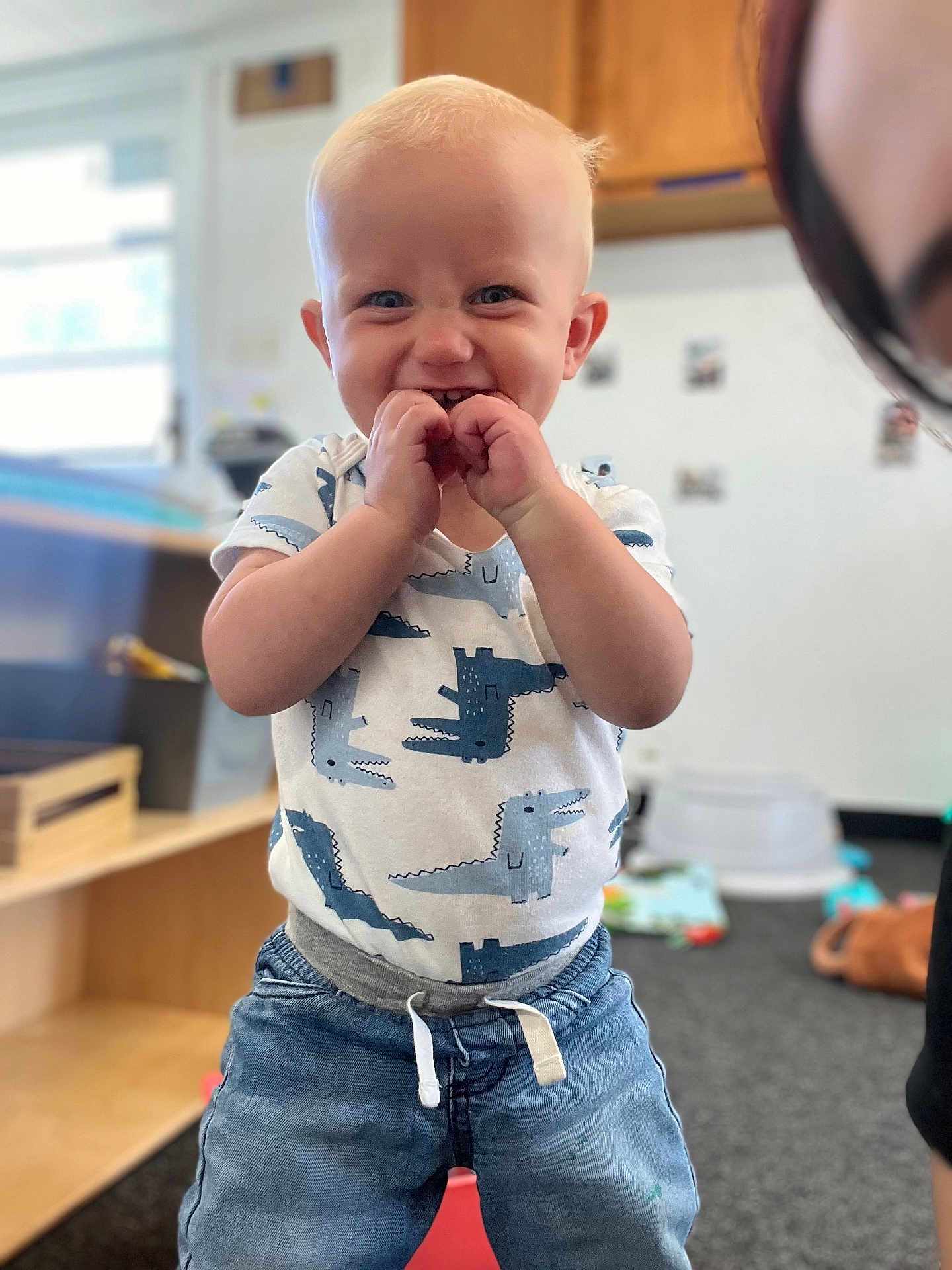 Hendrix is registered to the contest to win money with this photo: toddler, child, smiling, happy, indoor, clothing, dinosaur_print, shirt, denim_pants, standing, blue_eyes, blond_hair, playroom, carpet, toy, furniture, person, portrait, cute, young_child