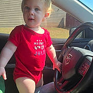 Elena is registered to the contest to win money with this photo: toddler, child, car_interior, steering_wheel, red_onesie, jeans, sunlight, window, seat, hand, face, blonde_hair, blue_eyes, person, clothing, casual, daylight, vehicle, driving, portrait