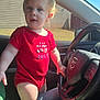 toddler, child, car_interior, steering_wheel, red_onesie, jeans, sunlight, window, seat, hand, face, blonde_hair, blue_eyes, person, clothing, casual, daylight, vehicle, driving, portrait