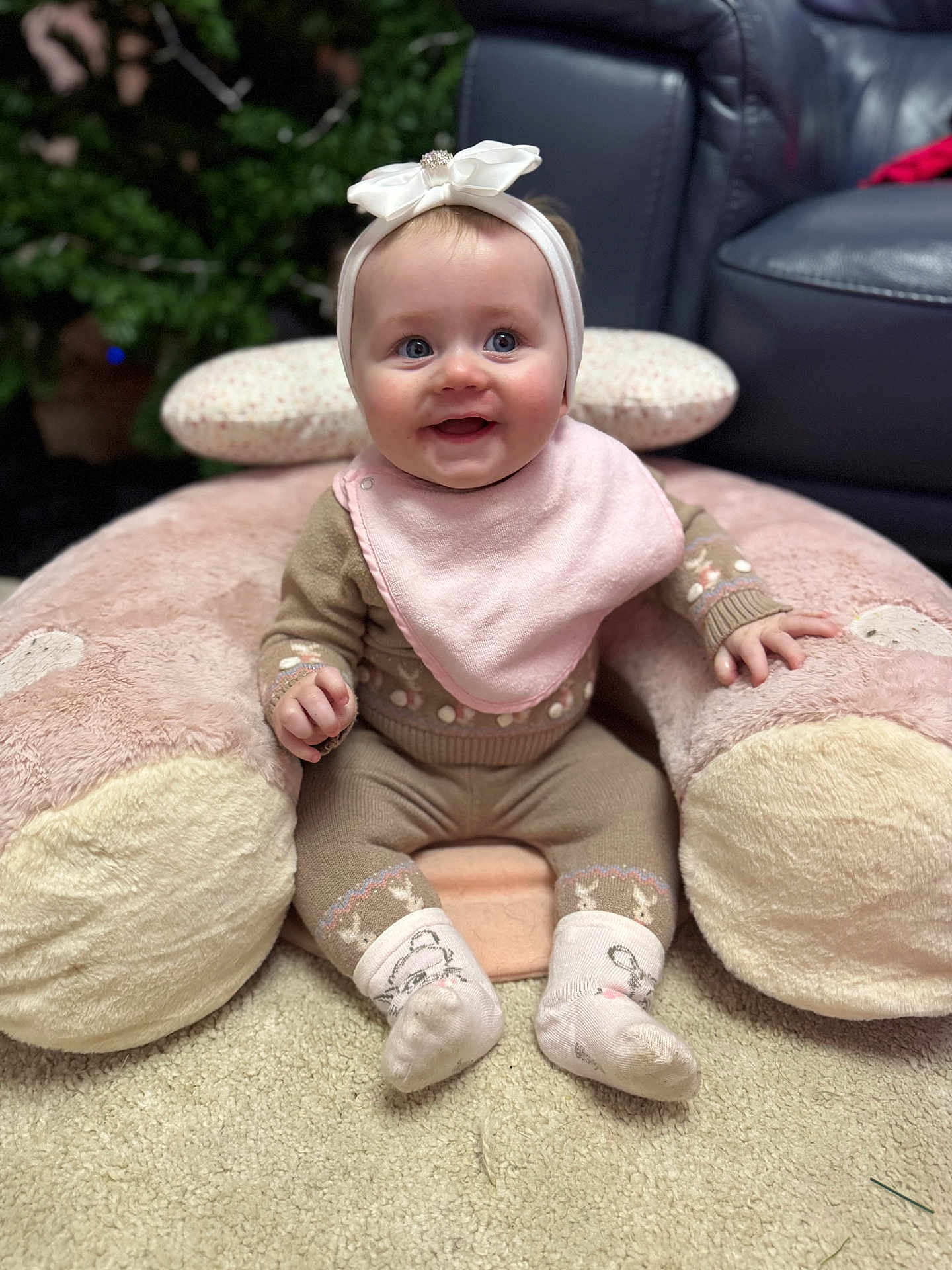 Blair Lilly is registered to the contest to win money with this photo: baby, headband, pink_bib, plush_pillow, carpet, socks, smile, indoor, child, cute, happy, seated, cozy, furniture, living_room, couch, warm_clothing, face, hands, blue_eyes
