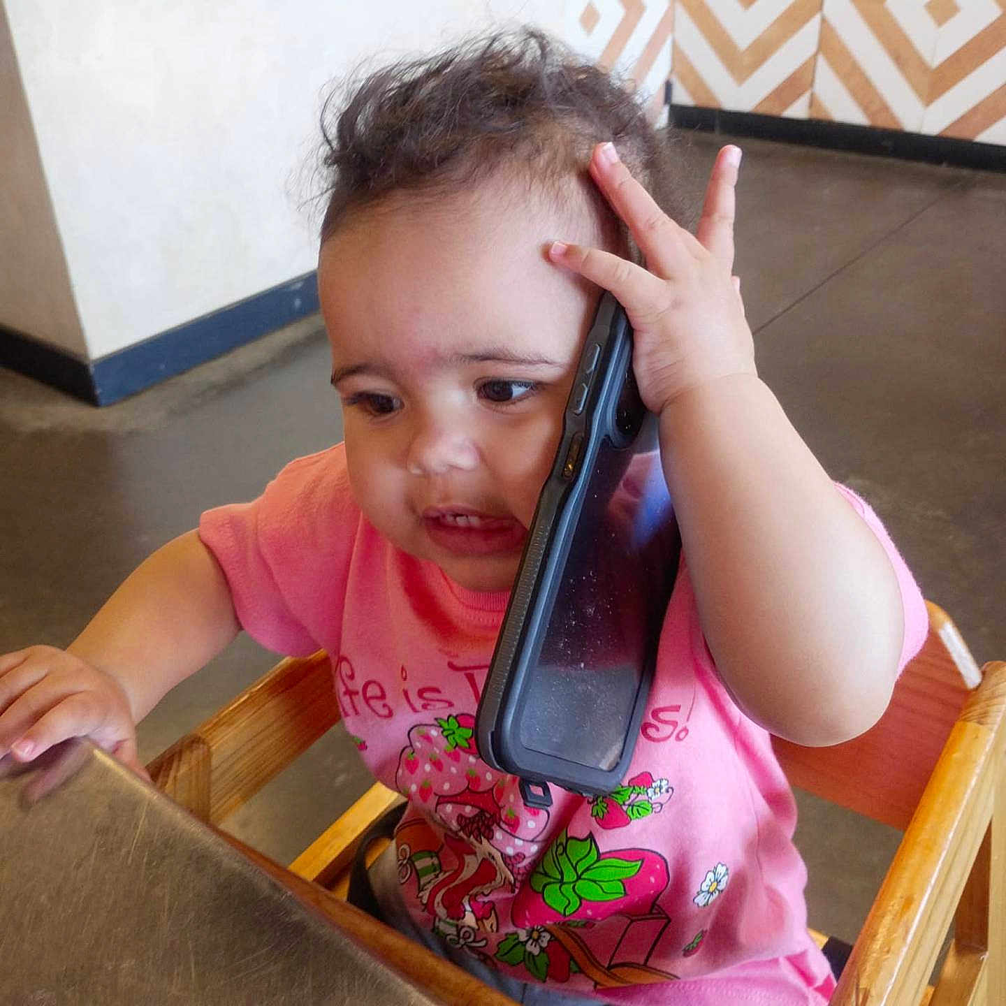 Lahlani joined the competition — help win amazing prizes! baby, cafe, child, curious, curly_hair, cute, face, hand, high_chair, indoor, phone_case, pink_shirt, portrait, sitting, smartphone, smiling, strawberry_print, table, toddler, wooden_chair