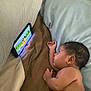 baby, child, tablet, cartoon, bed, blanket, pillow, indoor, screen, lying_down, curious, entertainment, digital_device, cozy, young_child, infant, watching, technology, relaxation, skin