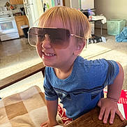 Oliver is registered to the contest to win money with this photo: blanket, blue_shirt, boy, cabinet, carpet, child, face, furniture, hair, hand, happy, home, indoor, kitchen, person, refrigerator, smile, sunglasses, table, wood