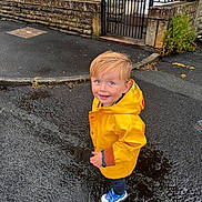 Hunter joined the competition — help win amazing prizes! building, child, curb, door, footwear, gate, greenery, happy, outdoor, person, puddle, raincoat, rainy_day, sidewalk, smiling, standing, stone_wall, weather, wet_street, yellow