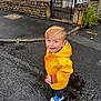 building, child, curb, door, footwear, gate, greenery, happy, outdoor, person, puddle, raincoat, rainy_day, sidewalk, smiling, standing, stone_wall, weather, wet_street, yellow