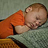 baby, blanket, child, closed_eyes, cozy, face, head, indoors, infant, knitted_blanket, lying_down, nap, newborn, orange_clothing, peaceful, portrait, resting, sleeping, soft_lighting, wooden_basket