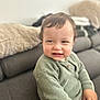 toddler, child, green_sweater, knitted_sweater, smile, cheeky_expression, couch, gray_couch, cushion, blanket, indoor, living_room, furniture, person, seated, short_hair, skin, hand, soft_lighting, casual_clothing