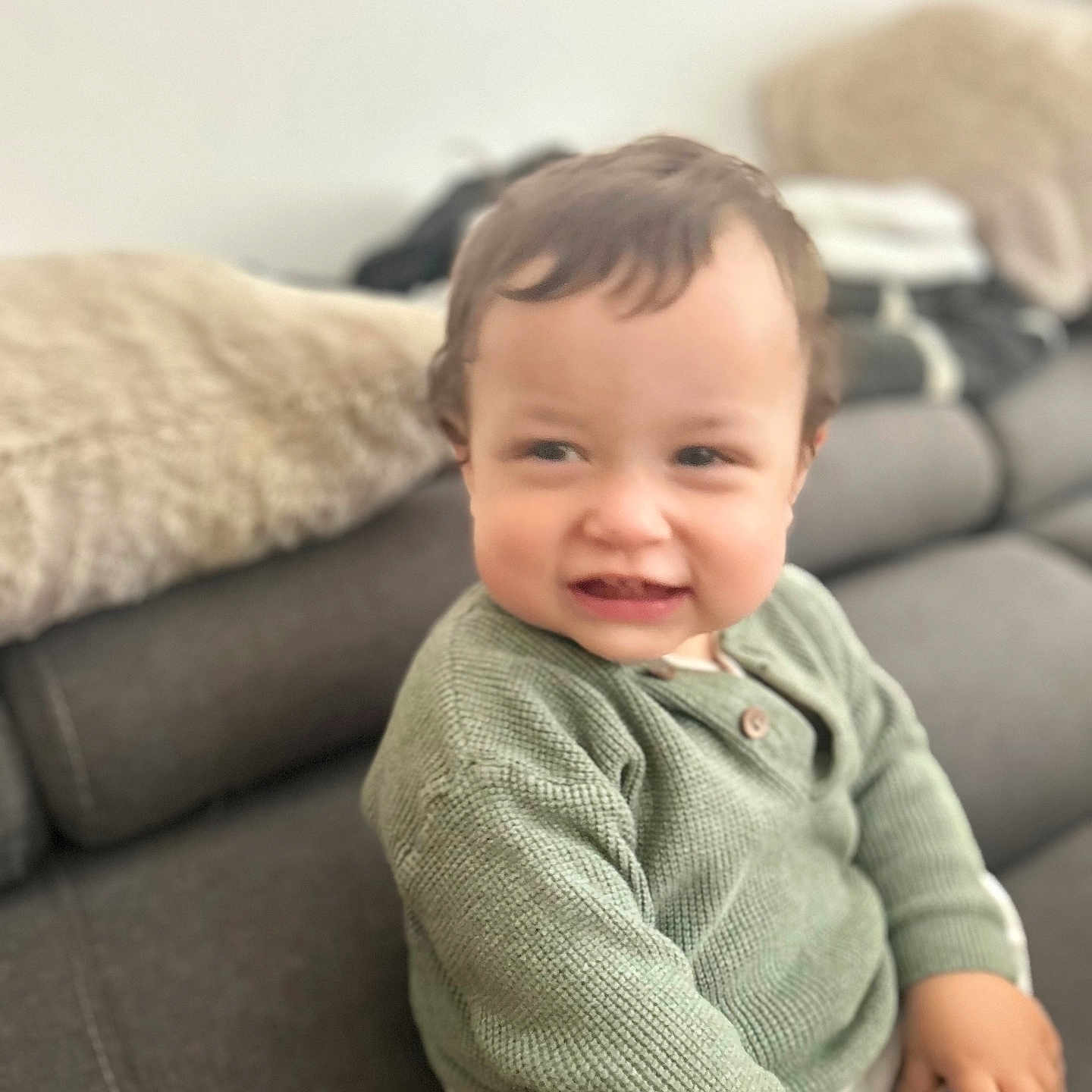 Isaac participe au concours pour gagner de l'argent avec cette photo : blanket, casual_clothing, cheeky_expression, child, couch, cushion, furniture, gray_couch, green_sweater, hand, indoor, knitted_sweater, living_room, person, seated, short_hair, skin, smile, soft_lighting, toddler