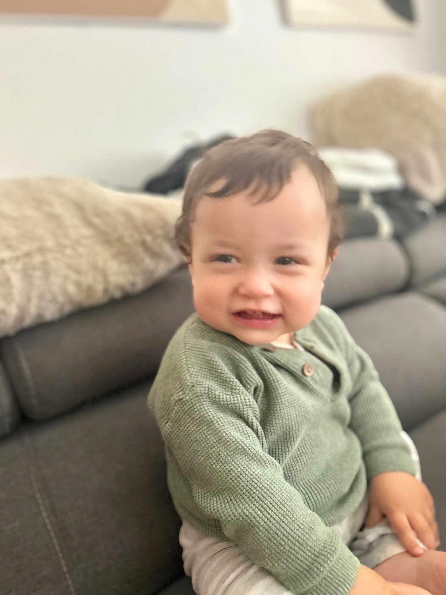 Isaac participe au concours pour gagner de l'argent avec cette photo : toddler, child, green_sweater, knitted_sweater, smile, cheeky_expression, couch, gray_couch, cushion, blanket, indoor, living_room, furniture, person, seated, short_hair, skin, hand, soft_lighting, casual_clothing