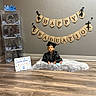 Douglas is registered to the contest to win money with this photo: baby, graduation, cap_and_gown, certificate, banner, burlap, flooring, wood_floor, rug, decorations, celebration, child, indoor, stacked_boxes, letters, blue_bows, black_clothing, sitting, portrait, smiling