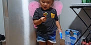 Kellcey is registered to the contest to win money with this photo: toddler, child, indoor, kitchen, refrigerator, lollipop, fairy_wings, shorts, sneakers, wood_floor, toy, curious, standing, person, hair_tie, plastic_bottle, box, table, shelf, happy