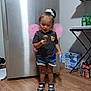 toddler, child, indoor, kitchen, refrigerator, lollipop, fairy_wings, shorts, sneakers, wood_floor, toy, curious, standing, person, hair_tie, plastic_bottle, box, table, shelf, happy