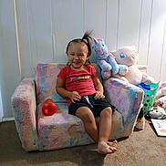 Kellcey joined the competition — help win amazing prizes! toddler, child, sofa, plush_toy, unicorn_toy, carpet, indoor, smiling, casual_clothing, red_shirt, black_shorts, barefoot, hair_ponytail, toy_bottle, laundry_basket, stuffed_animal, white_wall, happy, seated, home