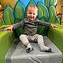 child, toddler, young_child, smiling, chair, green_throne, play_area, indoor, portrait, blond_hair, socks, long_sleeve_shirt, pants, hands, eyes, seat, happy, cute, mural_background, playground_prop