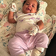 Khelani is registered to the contest to win money with this photo: baby, blanket, carpet, child, clothing, comfort, cute, face, floral, foot, headband, indoors, infant, lying_down, mittens, pattern, person, purple_pants, smile, soft_lighting