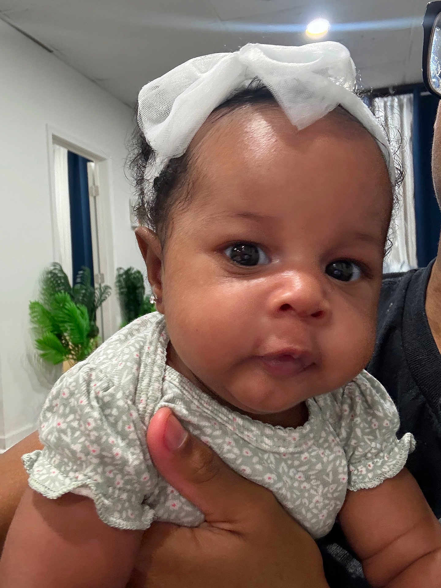 Khelani joined the competition — help win amazing prizes! baby, child, infant, face, headband, bow, hand, clothing, person, indoor, plant, doorway, curly_hair, cute, chubby_cheeks, portrait, holding, skin, expression, closeup