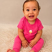 Alahia is registered to the contest to win money with this photo: baby, child, pink_clothing, sitting, smiling, happy, cute, indoor, portrait, fluffy_rug, barefoot, infant, looking_at_camera, short_hair, earrings, bright_colors, young_child, studio_lighting, isolated_background, playful