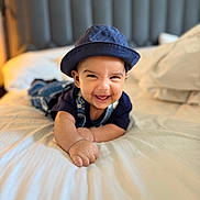 Zaydan is registered to the contest to win money with this photo: baby, bed, bedding, child, clothing, cozy, cute, denim_overalls, face, happy, hat, headwear, indoor, infant, person, portrait, room, skin, sleeping_area, smiling