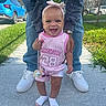 baby, child, smiling, standing, sidewalk, adult, hands, legs, pink_clothing, headband, outdoor, grass, car, shoes, happy, support, person, sunny, cute, portrait
