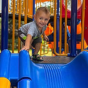 Randy is registered to the contest to win money with this photo: child, playground, slide, blue_slide, yellow_bars, orange_structure, smiling, short_hair, sandals, outdoor, daylight, metal_platform, clothing, happy, boy, play, fun, reaching, toy, park
