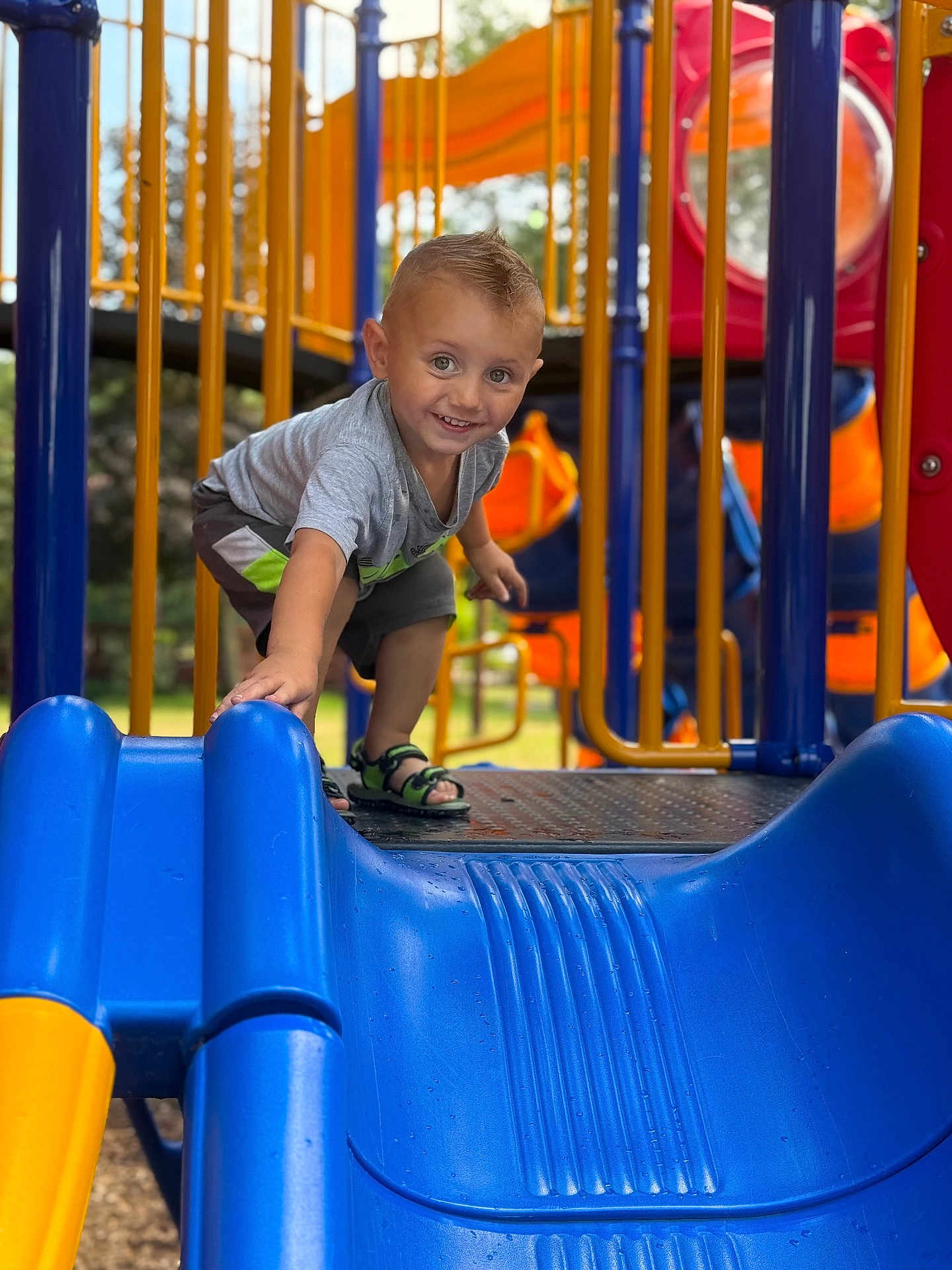 Randy is registered to the contest to win money with this photo: child, playground, slide, blue_slide, yellow_bars, orange_structure, smiling, short_hair, sandals, outdoor, daylight, metal_platform, clothing, happy, boy, play, fun, reaching, toy, park