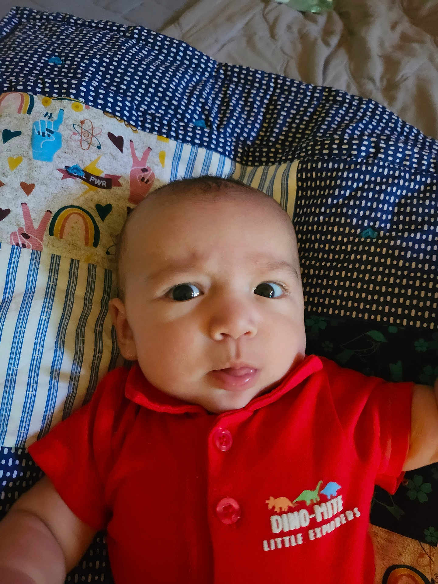 Amiri Lion joined the competition — help win amazing prizes! baby, infant, red_shirt, dinosaur_print, curious_expression, tongue_out, blanket, patterned_blanket, rainbow, peace_sign, closeup, face, person, child, indoors, cute, young, portrait, apparel, lying_down
