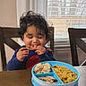 Noah is registered to the contest to win money with this photo: toddler, child, curly_hair, smiling, eating, food, plate, blue_plate, table, chair, window, blinds, indoor, meal, happy, person, cute, curtains, wooden_table, casual