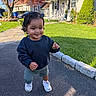 toddler, child, curly_hair, sneakers, sweatshirt, shorts, sidewalk, driveway, grass, tree, house, daylight, smiling, outdoor, suburban, person, young_child, walking, shadow, nature