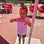 child, girl, outdoor, sunny, heart_shape, white_shirt, black_shorts, sneakers, red_chairs, table, umbrella, fence, bracelet, hair_tie, pavement, playground, daylight, person, casual_wear, smiling