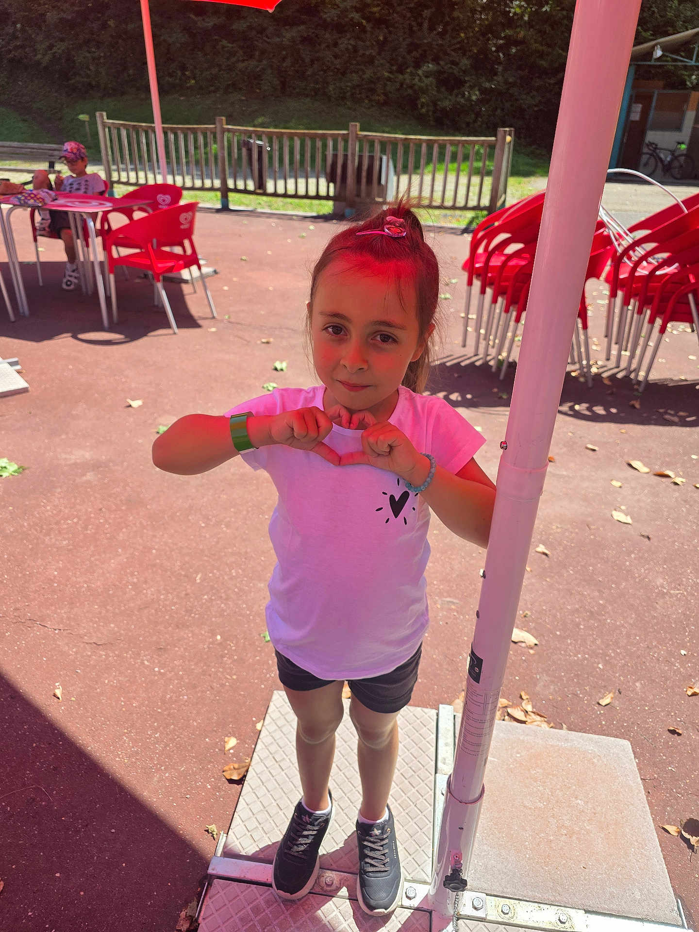 Myléna participe au concours pour gagner de l'argent avec cette photo : child, girl, outdoor, sunny, heart_shape, white_shirt, black_shorts, sneakers, red_chairs, table, umbrella, fence, bracelet, hair_tie, pavement, playground, daylight, person, casual_wear, smiling