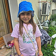 Myléna participe au concours pour gagner de l'argent avec cette photo : bag, blue_hat, bracelet, casual_clothing, chair, child, concrete, garden, girl, greenery, horse_print, house_wall, long_hair, outdoor, pink_tshirt, plants, smiling, towel, toys, window
