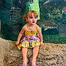child, toddler, girl, portrait, aquarium, fish, underwater, colorful_dress, sitting, rock, outdoor, indoor, wildlife, marine_life, glass_tank, curious, brown_hair, smiling, sandals, family_photo