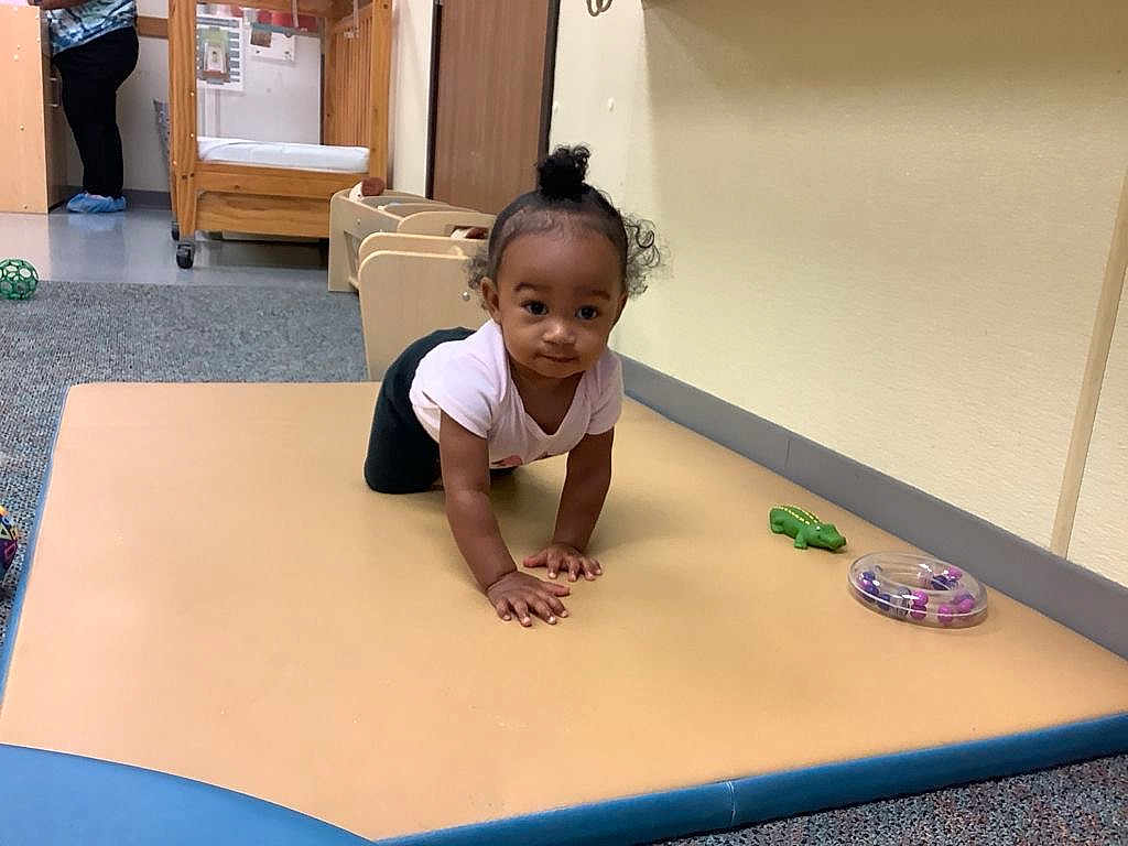 Autumn is registered to the contest to win money with this photo: toddler, child, crawling, mat, toy, playroom, carpet, wooden_crib, person, shoe_covers, topknot, curly_hair, indoor, floor, wall, casual_clothing, baby, young_child, play, curious
